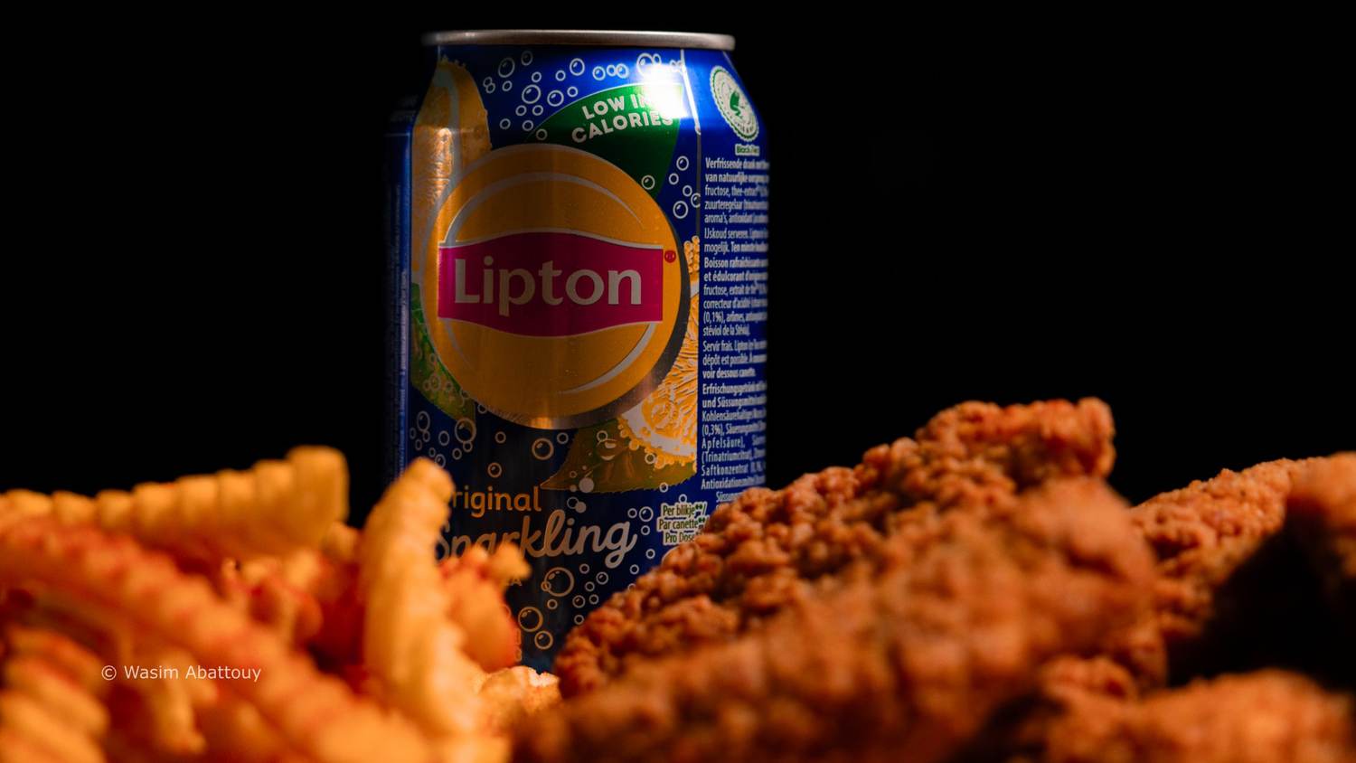 An iced tea can behind fries and fried chicken, lit against black