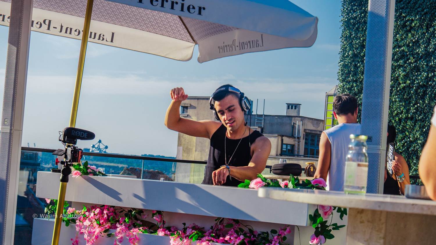DJ with an arm raised behind a booth covered in pink flowers, under parasols
