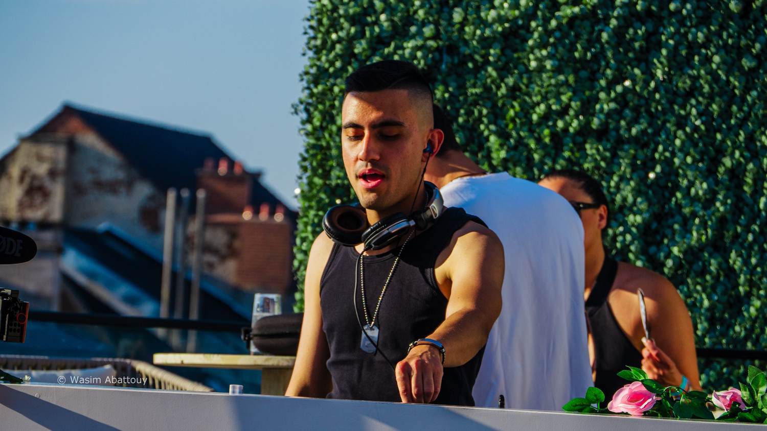 DJ with headphones around his neck, concentrating, in front of a green hedge wall