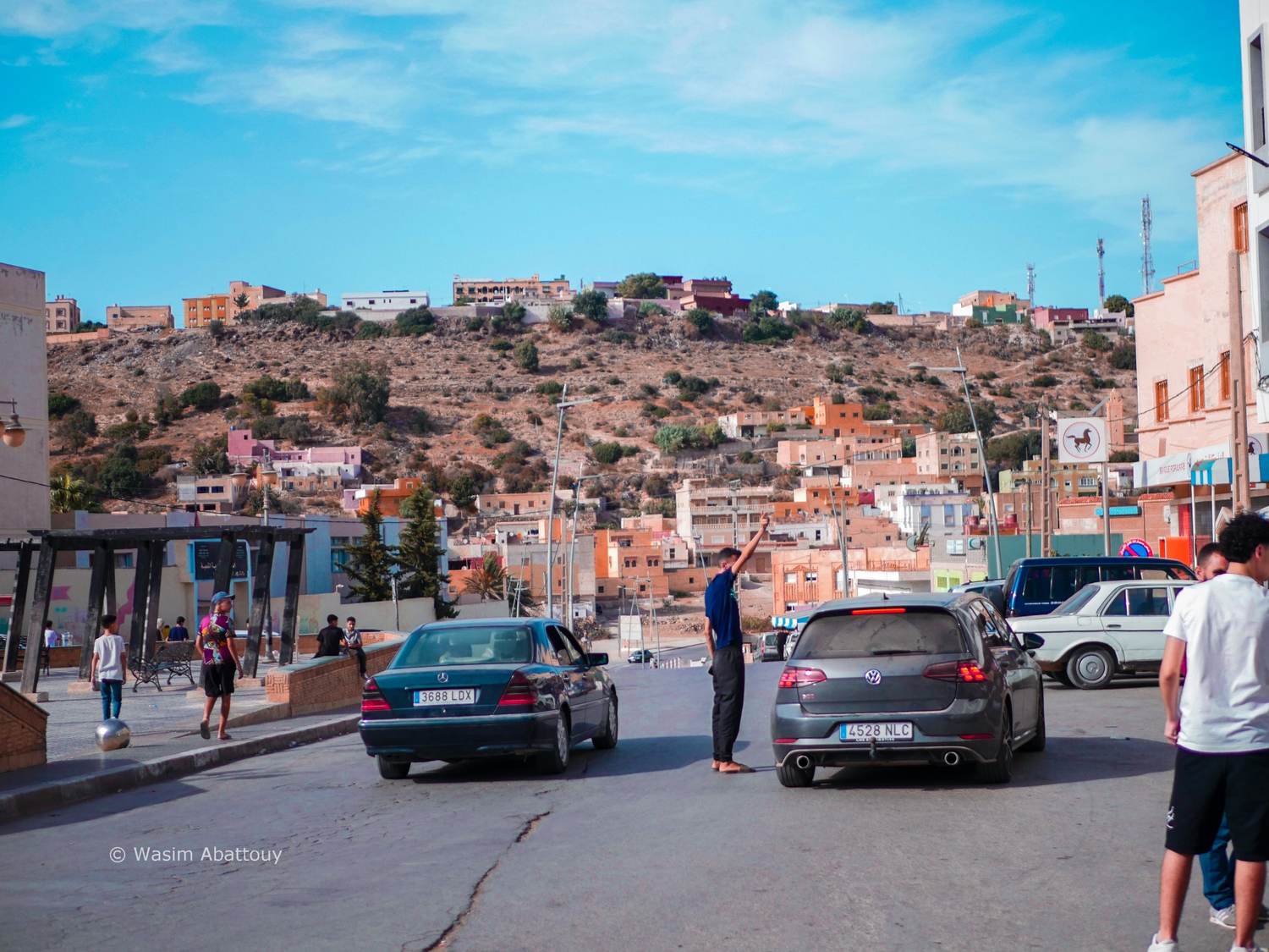 A hillside town with pastel houses above a busy street, cars and people in the foreground