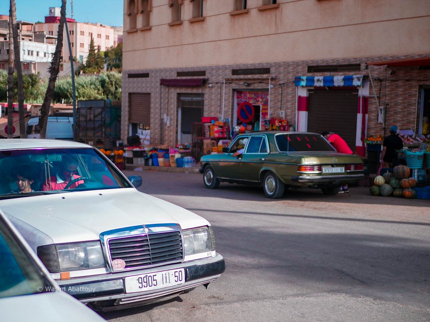 A white taxi and an old green Mercedes parked in front of a fruit and vegetable shop
