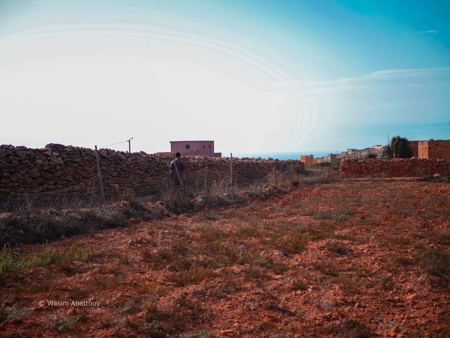 A person walking along a dry stone wall and fence across red earth, with the sea on the horizon